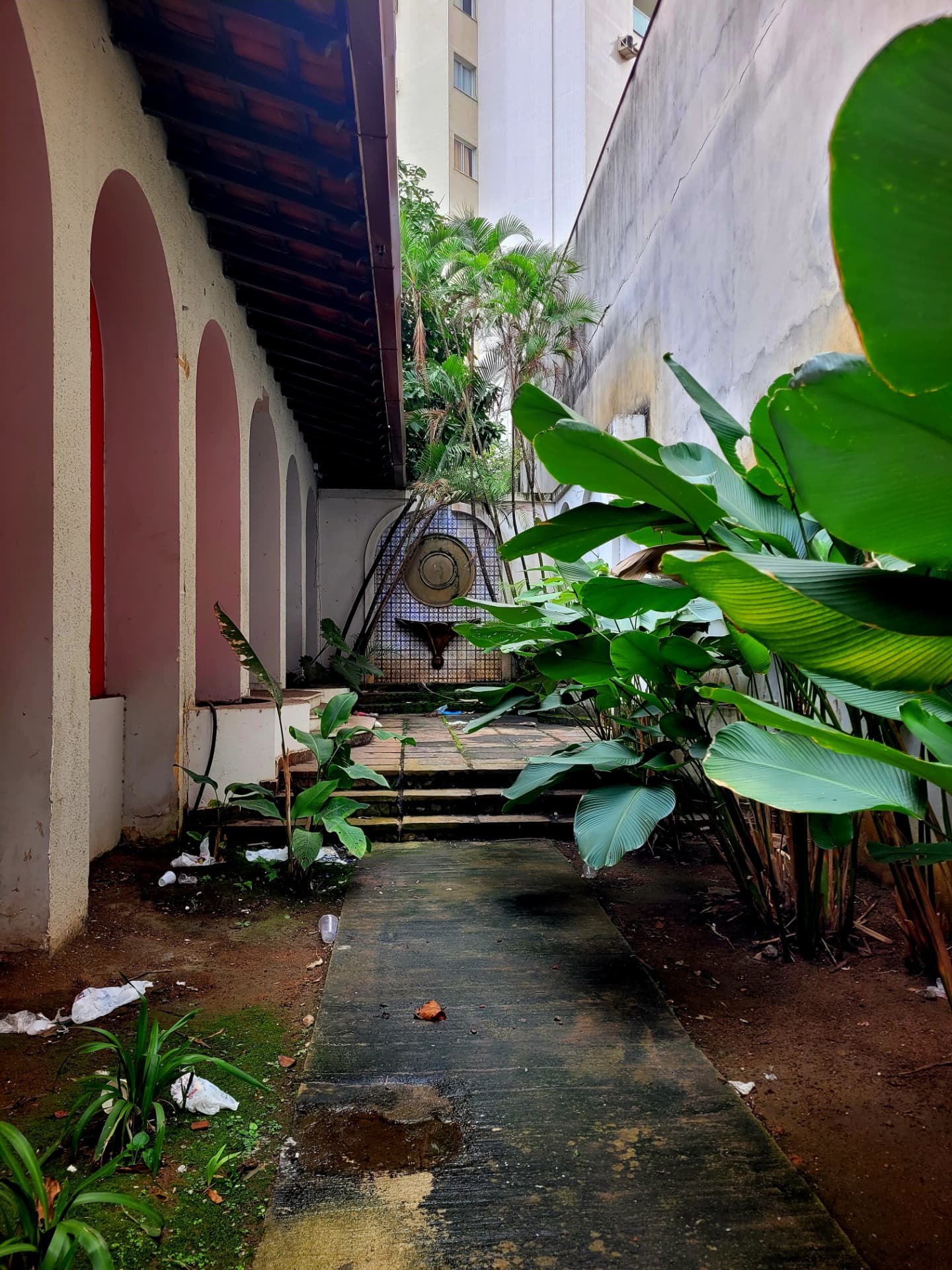 Corredor de uma casa antiga escondida entre prédios, com arcos, janelas vermelhas, jardim tomado por plantas e uma fonte ao fundo